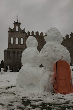 A lively winter scene at the northern stone fortifications of Avila, Spain, where locals and visitors play in the deep snow and build snowmen against the backdrop of the historic medieval walls.