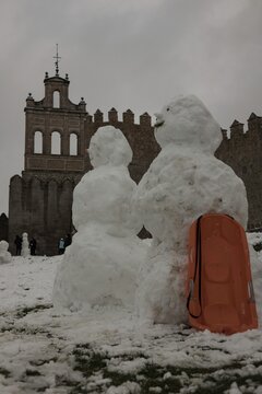 A lively winter scene at the northern stone fortifications of Avila, Spain, where locals and visitors play in the deep snow and build snowmen against the backdrop of the historic medieval walls.