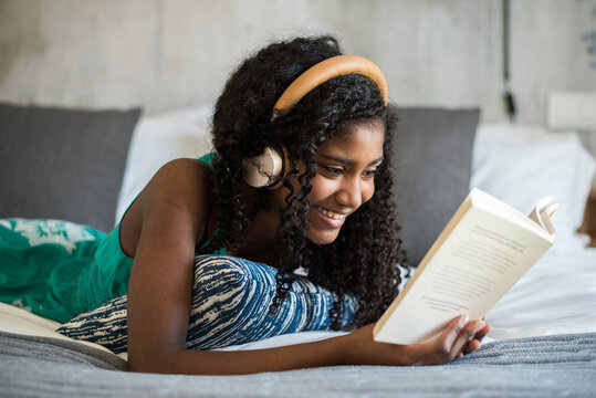 Black teenager reading a book on bed with headphones