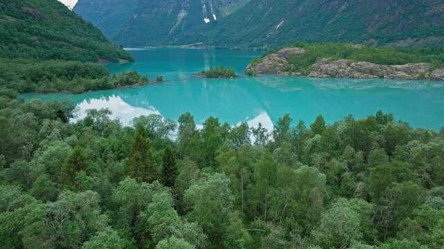 Aerial view over bright turquoise Lovatnet lake in Norway. Forested foreground and alpine mountains frame the glacial water on a serene summer day.