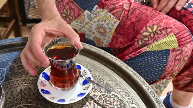 Istanbul,turkey,august 6, 2025. Close-up of a woman's hand picking up a traditional tulip-shaped glass of turkish tea from an ornate metal tray in a classic cafe