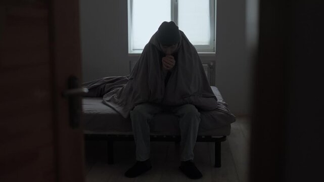 No heating at home, man sitting under blanket in cold room, winter energy crisis lifestyle.