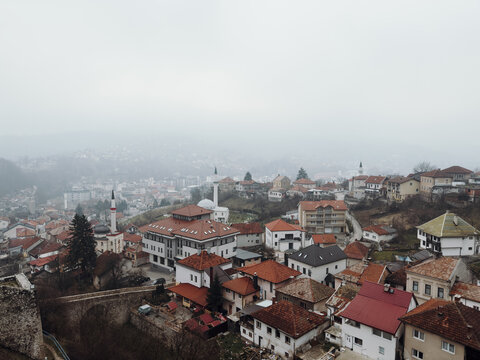 Misty panoramic view of Travnik with dense residential buildings spreading across hills, softened by fog and muted winter colors, emphasizing depth and atmospheric urban scenery.