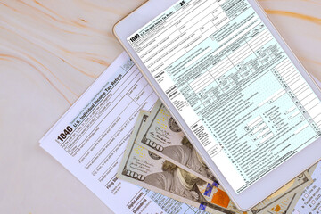 Obraz premium Digital tablet displays tax 1040 forms while US cash banknote is scattered on table with other tax documents.