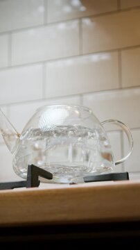 Transparent glass teapot sits on a gas burner in a tiled kitchen, warming up before tea preparation begins, vertical video