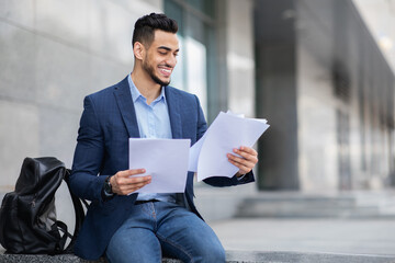 Excited young arab man in formal outfit sitting on the street next to business building, holding documents or CV, having backpack by him, getting ready for job interview, panorama with copy space