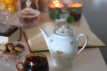 Cup of tea or coffee, teapot, book, notebook, e-reader, glasses, cookies, nuts, lemons, house plants and lit candles on the table. Hygge at home. Selective focus.