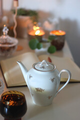 Cup of tea or coffee, teapot, book, notebook, e-reader, glasses, cookies, nuts, lemons, house plants and lit candles on the table. Hygge at home. Selective focus.