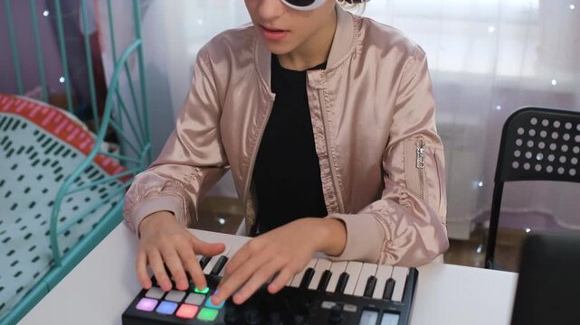 Girl enjoys music by crafting sounds on a midi keyboard while wearing headphones and sunglasses, showing her love for creating in her home space