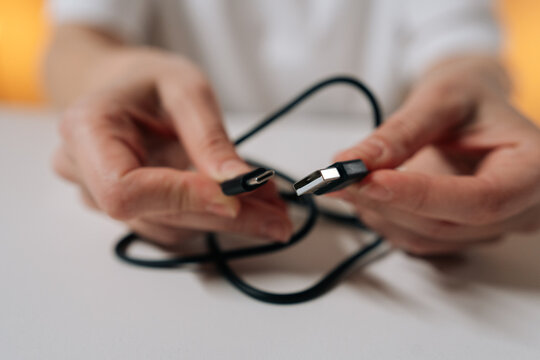 Close-up hands of unrecognizable person untangling knotted black power cord sitting at white table, preparing charging cable for use and restoring order to tangled wire.