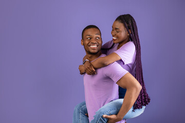 Obraz premium Piggyback Ride. Smiling black man carrying and riding his woman with long afro braids on back, enjoying time together, standing isolated on violet studio background, having fun, free copy space