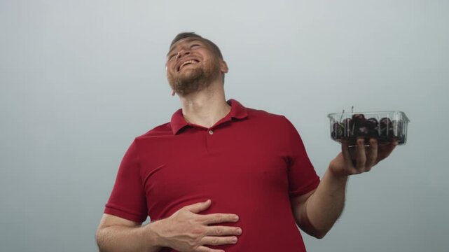 Man smiling and holding plastic container of cherries in studio with pale gray background; joy healthy snack.