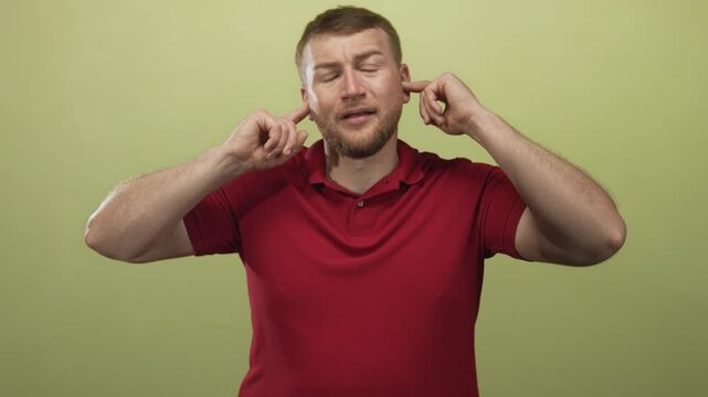 Man in red polo shirt plugging fingers into his ears in a light green studio, wincing and closing eyes; annoyance avoidance.