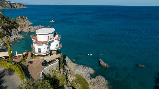 Aerial view of a unique, multi-story white circular villa perched on a rocky cliff overlooking a vast, clear turquoise sea, and a lush Mediterranean garden surrounding the property.