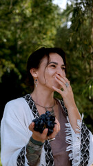 A beautiful brunette in a bohemian style relaxes outdoors in summer against a backdrop of a green forest, holding a large bunch of juicy black grapes