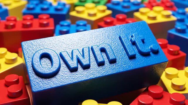Blue Lego brick with the phrase Own It on top of multicolored Lego bricks from a close-up viewpoint