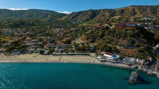 Aerial view of a coastal village nestled at the foot of lush, rolling hills, featuring a sandy beach that meets clear turquoise waters. It is the Caminia waterfront, near Soverato in Calabria, Italy.