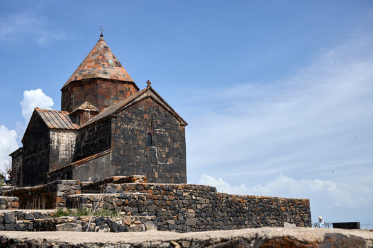 Ancient stone church with a conical roof under a clear blue sky, surrounded by a stone wall. The structure features dark, weathered stones and a serene, open atmosphere.