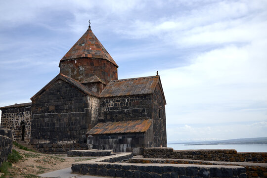 Ancient stone church with a conical roof, set against a cloudy sky and distant mountains. The structure features dark, weathered stones and overlooks a serene body of water.