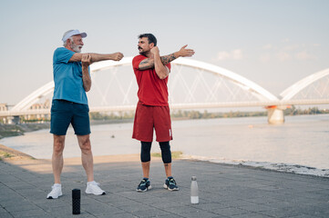 Fototapeta premium Senior man and personal trainer stretching arms by river