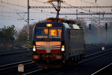 Photo of an electric train approaching on railway tracks at dusk. Headlights, overhead lines, and rails emphasize modern rail transport, motion, and infrastructure.