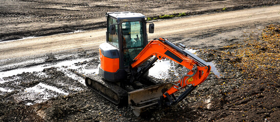 Mini excavator digging soil at construction site near road. Heavy machinery in action, earthmoving process, building infrastructure and engineering work concept. © VladyslavShcherbakov
