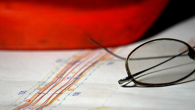 Eyeglasses resting on technical blueprint with colorful graph lines and blurred red background. Concept of engineering, planning, analysis and professional design work.