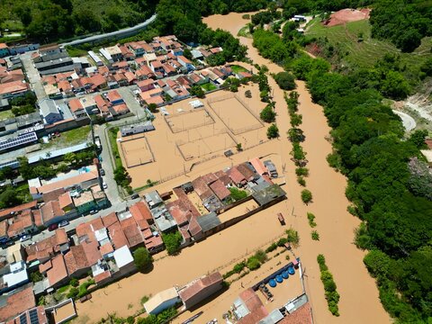 Vista a&eacute;rea por drone de uma enchente urbana, mostrando ruas alagadas, casas parcialmente submersas e o impacto das chuvas intensas. Conceito de desastre natural, mudan&ccedil;as clim&aacute;ticas, crise ambiental.