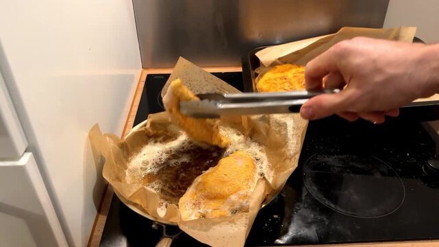 A man's hand flipping meat in a frying pan. Hand holding tongs. Frying breaded chicken. Home cooking.
