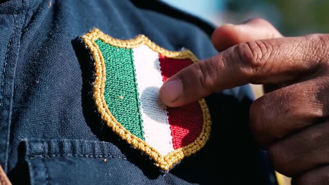 Close up shot featuring a person's finger gently touching a small embroidered shield patch displaying the vibrant colors of the Italian flag sewn onto dark blue fabric