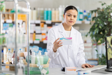 Fototapeta premium Positive middle-aged woman apothecary showing medicament standing by computer in drugstore