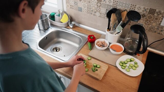 Over the shoulder view of teenage boy cut fresh green cucumber on wooden board while prepare healthy vegetable salad in modern cozy home kitchen