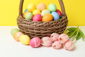 Painted Easter eggs in wicker basket and beautiful tulips on white table against yellow background, closeup