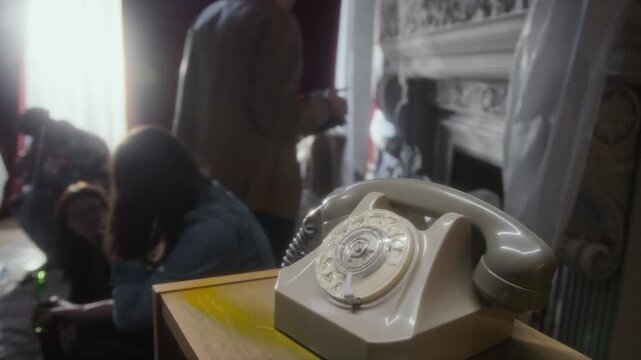 Handheld shot of old rotary phone on stand with group of young people hanging out together in abandoned house together in background, copy space