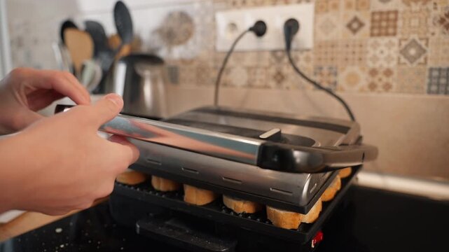 Person carefully places slices of bread onto hot electric grill, then closes lid to toast them into crispy croutons, perfect for delicious homemade breakfast or party appetizer