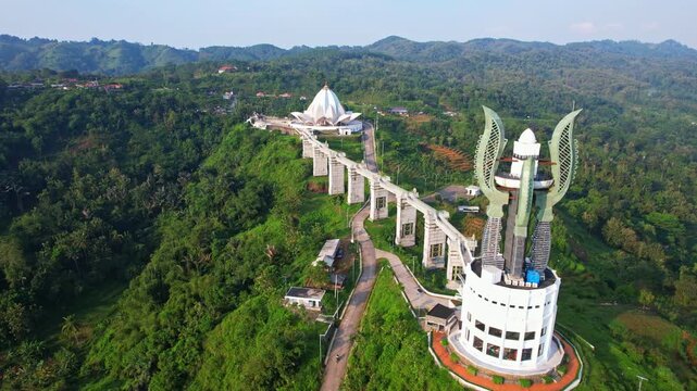 Aerial drone footage of Kujang tower, with a bridge leading to the lotus flower shape Al Kamil beautiful mosque, a road, at sunset, mountains behind, in Jatigede reservoir, Sumedang regency, Indonesia