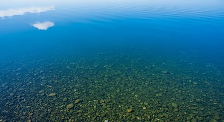 Fototapeta premium Crystal clear blue water reveals smooth pebbles on the lakebed under a bright sky