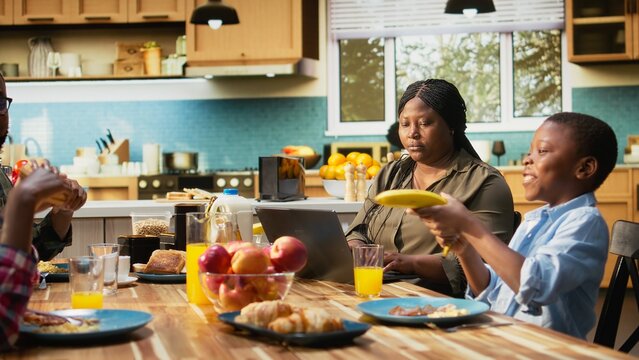 African American mom does remote work on laptop while the kids play with food, parents and children eating a homemade meal together in the kitchen. Goofy children fight at table. Camera A.