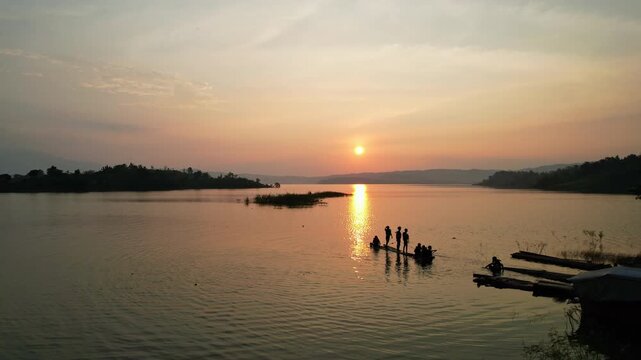 Aerial drone footage at sunset of a small wooden skiff, with few people on it as silhouettes, in flat waters, in Jatigede reservoir, with islands and sun behind, in Sumedang regency, Indonesia