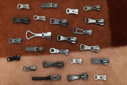 A collection of various metal zipper pulls with different shapes and finishes is scattered across a textured brown surface.
