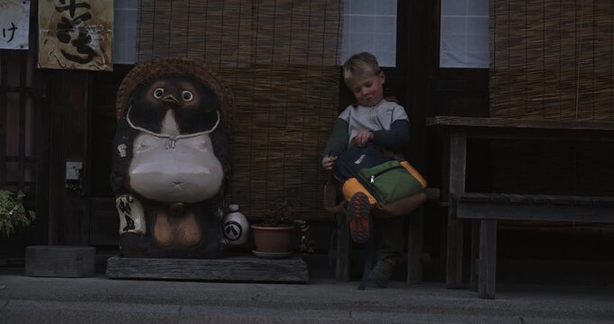 Little boy sitting in front of Japanese structure near bear statue and opening backpack