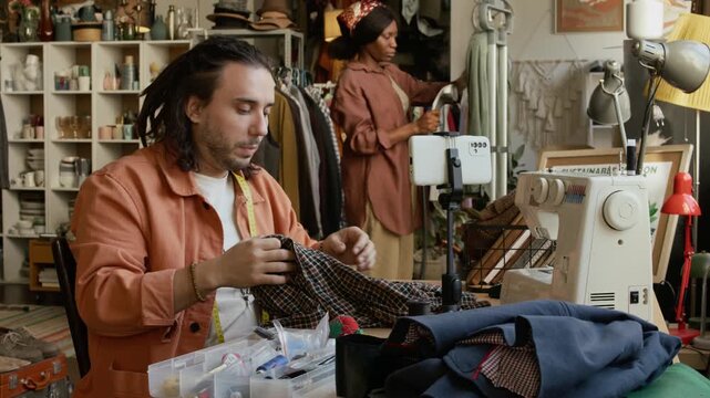 Medium shot of young Caucasian male second hand shop owner sitting at table with sewing machine, setting up smartphone then recording tutorial on upcycling old clothes for social media blog