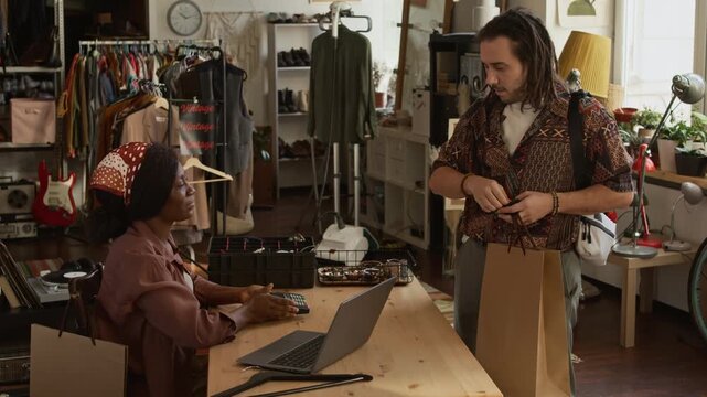 Medium shot of smiling young Caucasian man taking shopping bag from black female assistant while making purchase at second hand outlet, paying with credit card, happy with bargain