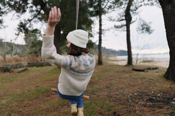 Outdoor swing in a forested park with a child wearing a knit sweater and beanie, swinging gently by a rope with distant lake and hills creating a tranquil nature scene