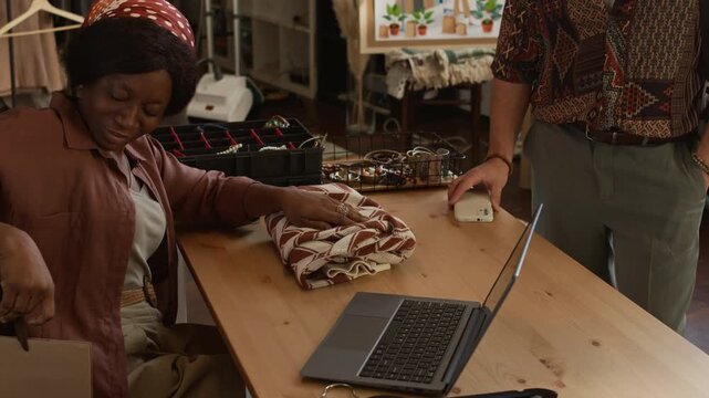 Medium shot of young African American sales assistant serving male customer at second hand store, folding patterned vintage sweater, packing in shopping bag, man paying for purchase with smartphone