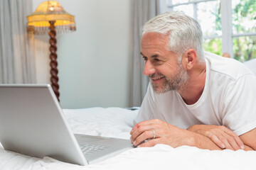 Fototapeta premium Senior man lying on bed propped on elbows, looking at silver laptop and wearing wedding band