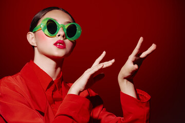 Fototapeta premium Fashion model with bold green eyewear and bright red lipstick poses with expressive hands wearing a matching red outfit against a red background. Creative beauty concept.
