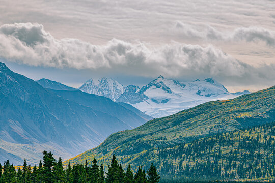 Snow-covered peaks rise above rolling tundra hills and boreal forest under dramatic clouds along the road from Fairbanks to Valdez, Alaska.