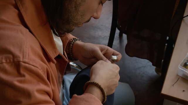 Medium high-angle shot of crafty young Caucasian man with dreadlocks sewing elbow patch onto preowned jacket in creative workshop, while creating upcycled clothing for sustainable lifestyle
