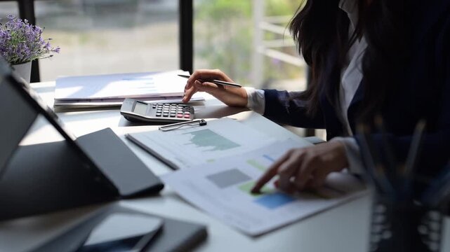 A woman is sitting at a desk with a calculator and a piece of paper with graphs on it. She is pointing at the paper and she is focused on the numbers